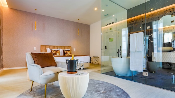 Une chambre d’hôtel moderne équipée d’un lit, d’un canapé, d’une petite table avec une boisson, et d’une salle de bain fermée par des parois de verre.