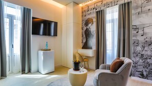 Un salon moderne avec un téléviseur à écran plat fixé au mur, un mini-réfrigérateur blanc, une petite table ronde avec une bouteille de champagne et des verres, un canapé gris avec un coussin marron et un mur orné d’un motif de notes de musique.