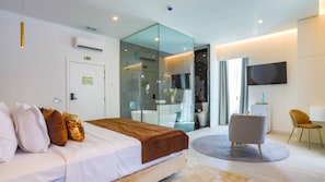 Une chambre d’hôtel moderne équipée d’un lit, d’un coin salon, d’une télévision et d’une salle de bain fermée par des parois de verre.