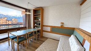 A room with a dining table, chairs, a bookshelf, a bed, and a view of mountains. - Apartment '- Les 2 Alpes' with Mountain View, Shared Pool and Wi-Fi (Les Deux Alpes)