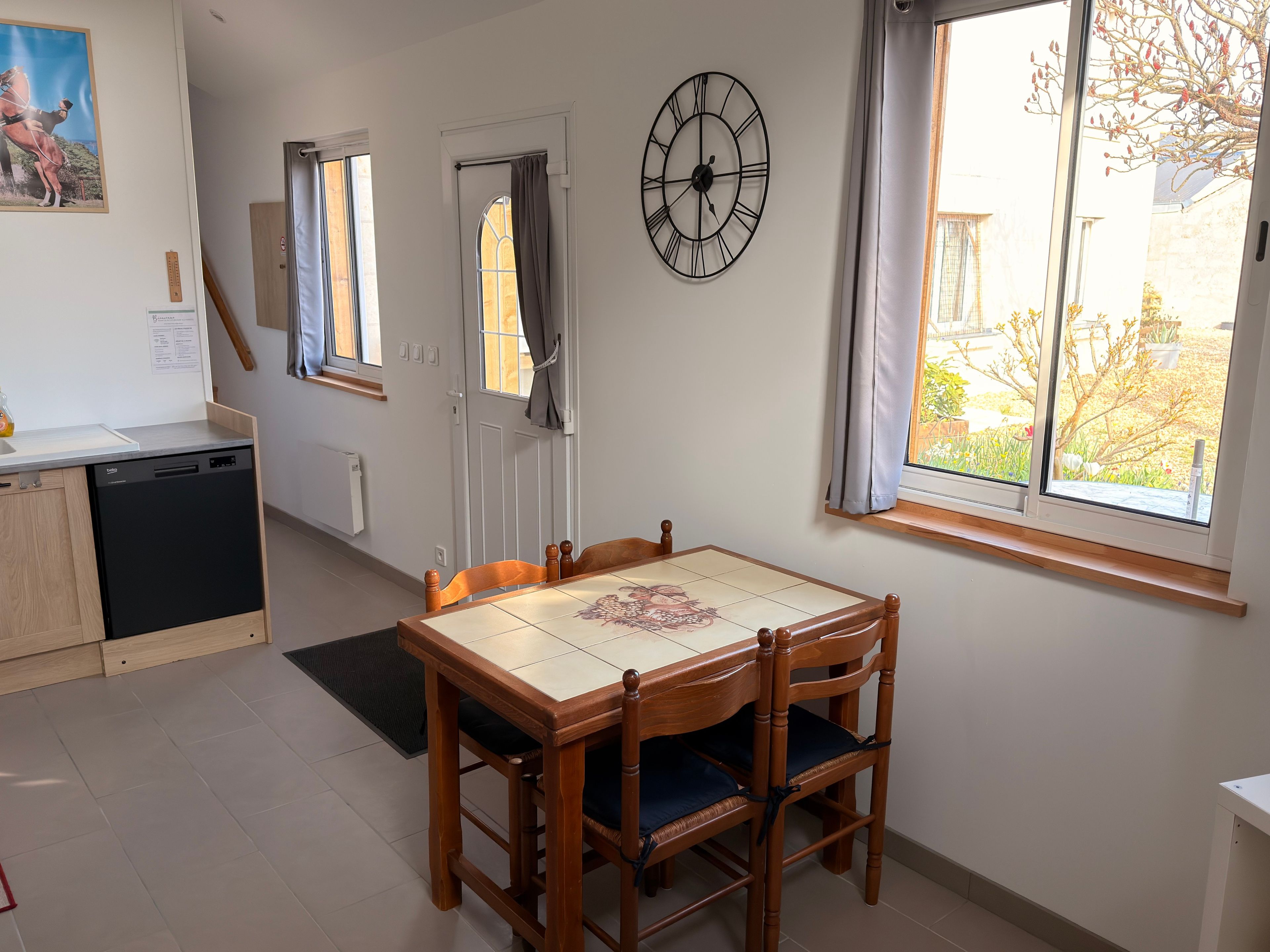 Una zona de menjador amb taula i cadires de fusta, un rellotge a la paret i una finestra amb cortines.