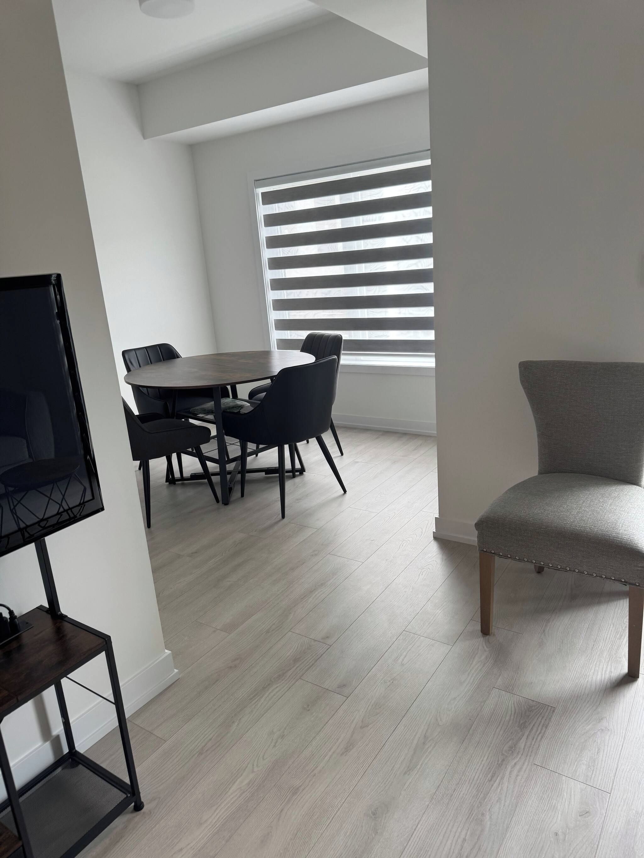 Uma área de refeições moderna com uma mesa redonda e cadeiras, uma televisão em um móvel e uma janela com persianas.