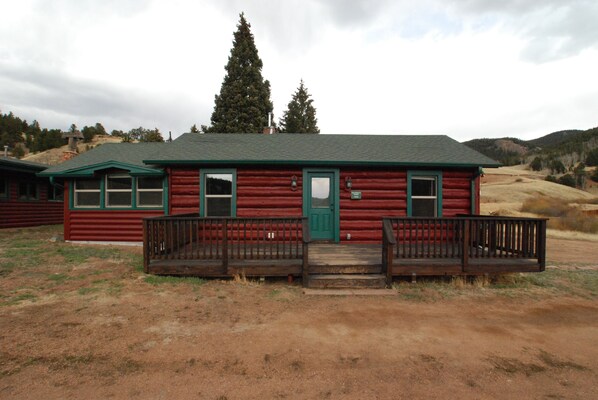 A red log cabin with a green door, two windows, and a porch with a bench.