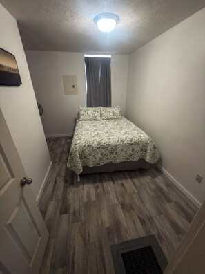 A small, single-room apartment with a bed, a window with curtains, a door, and a ceiling light. - 1BR close to the Erie Canal Trail, and across from Lunkenheimer Craft Brewery (Weedsport)