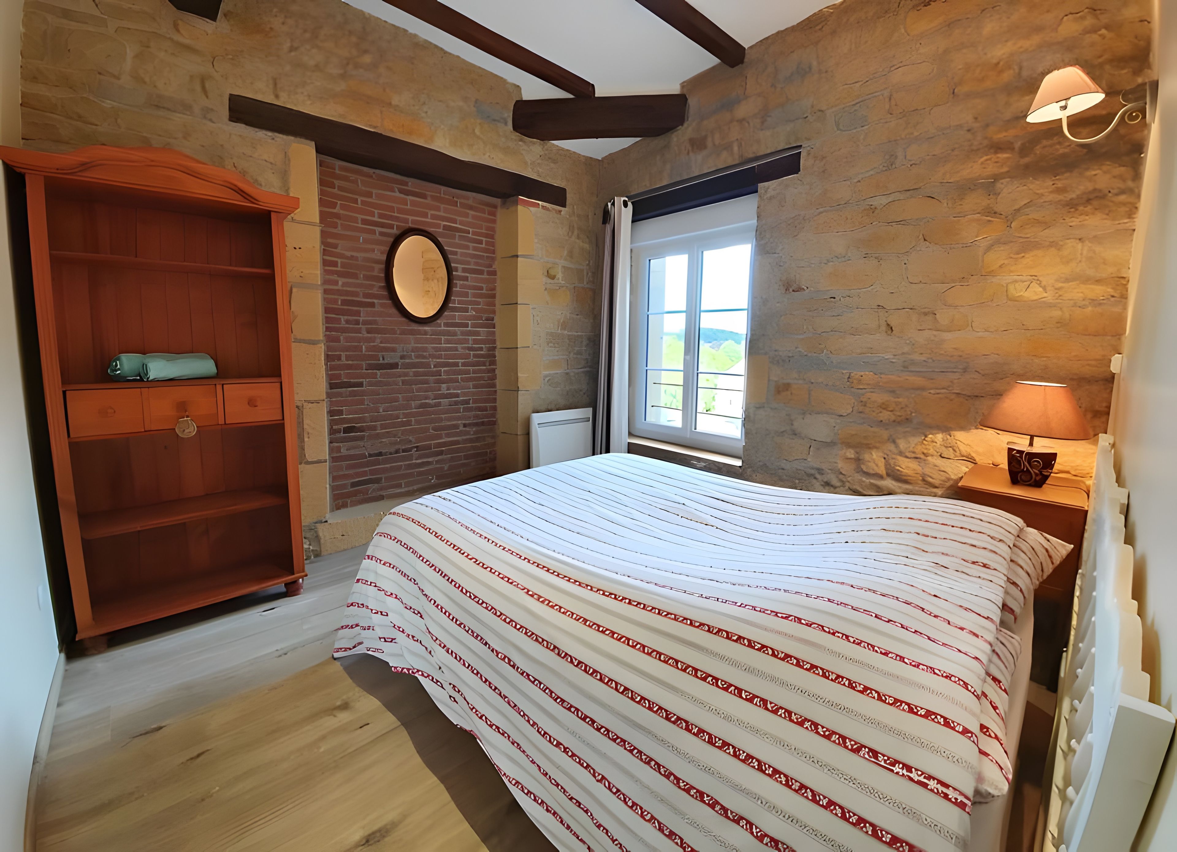 Een slaapkamer met een houten garderobe, een bed met gestreepte lakens, een stenen muur en een raam met uitzicht.