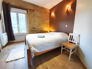 Een slaapkamer met een bed, nachtkastje, stoel, raam en een wandlamp.