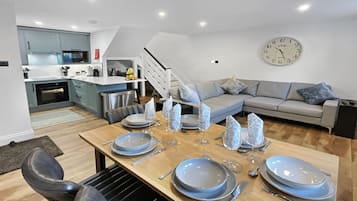 Un espai d'estar modern amb zona de dinar preparada per a sis, cuina amb armaris grisos i escala que condueix a un pis superior.