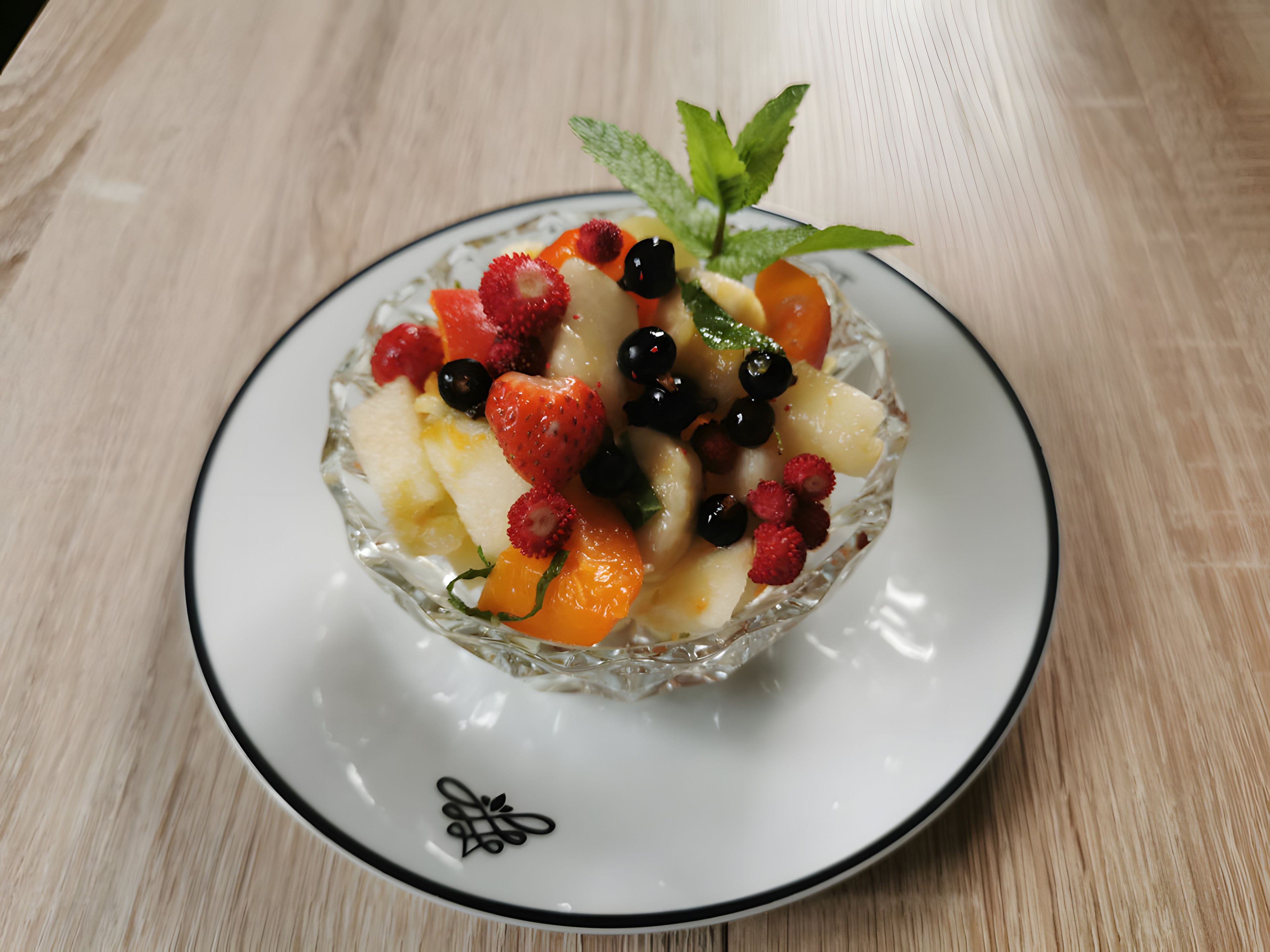 A clear glass bowl filled with assorted fruits on a white plate with a black rim, placed on a wooden surface.