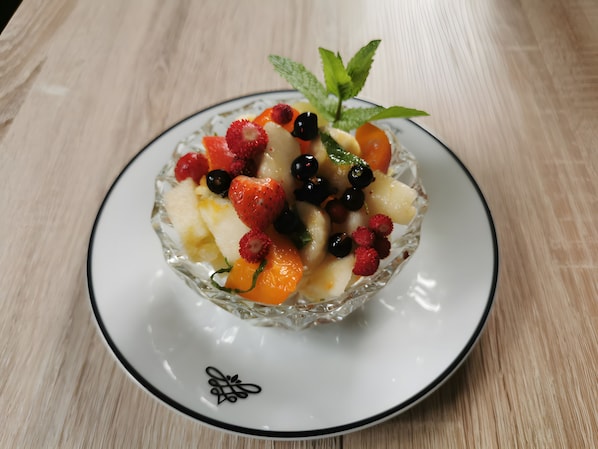 Un saladier en verre transparent, rempli d’un assortiment de fruits, est posé sur une assiette blanche à bord noir, elle-même placée sur une surface en bois.