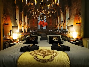 Une chambre d’hôtel avec un grand lit, deux oreillers ornés d’un blason, une table de chevet avec une lampe, et une vue sur un vitrail.