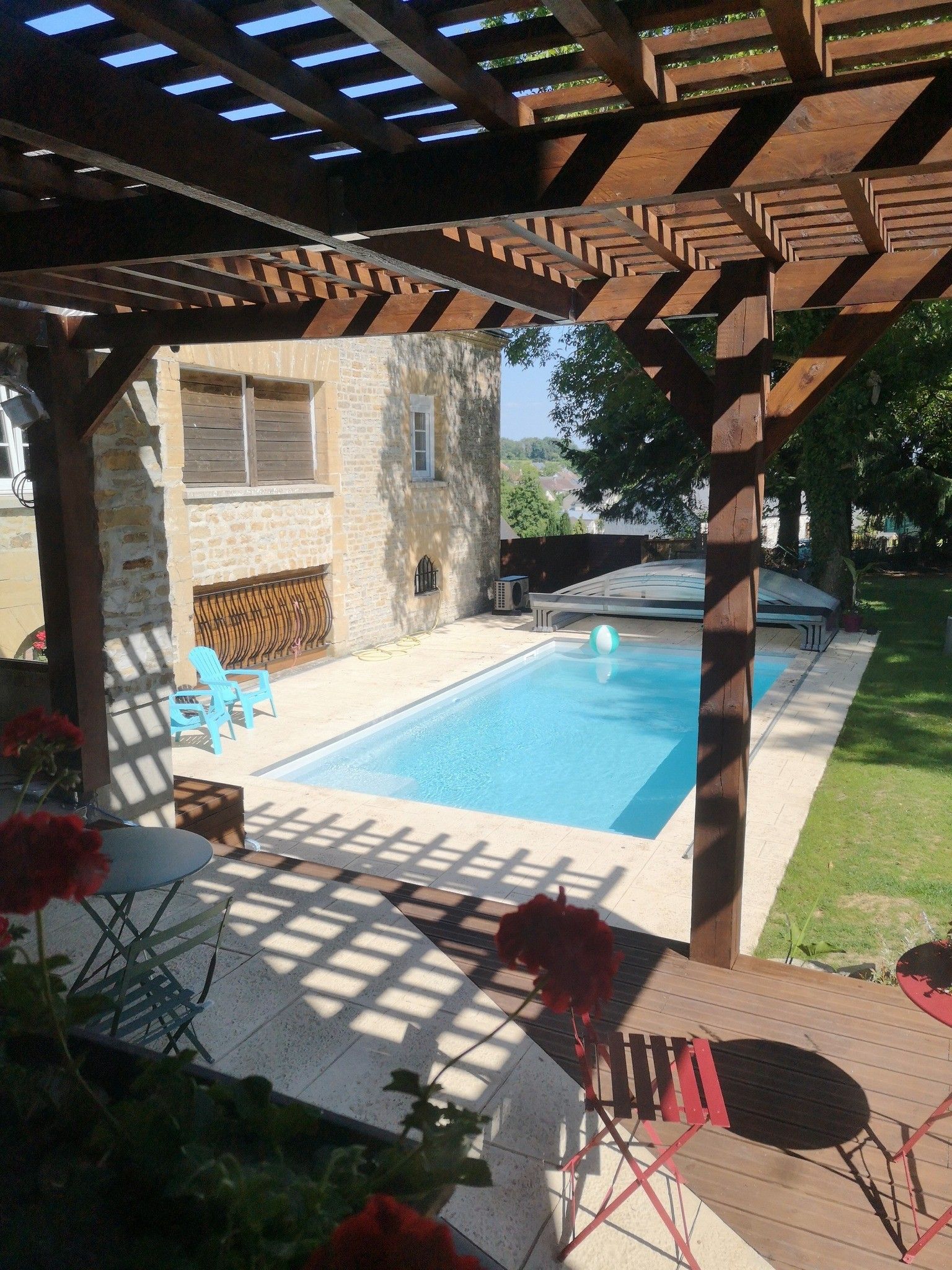 A covered outdoor swimming pool with a stone wall, wooden deck, and a pergola overhead.