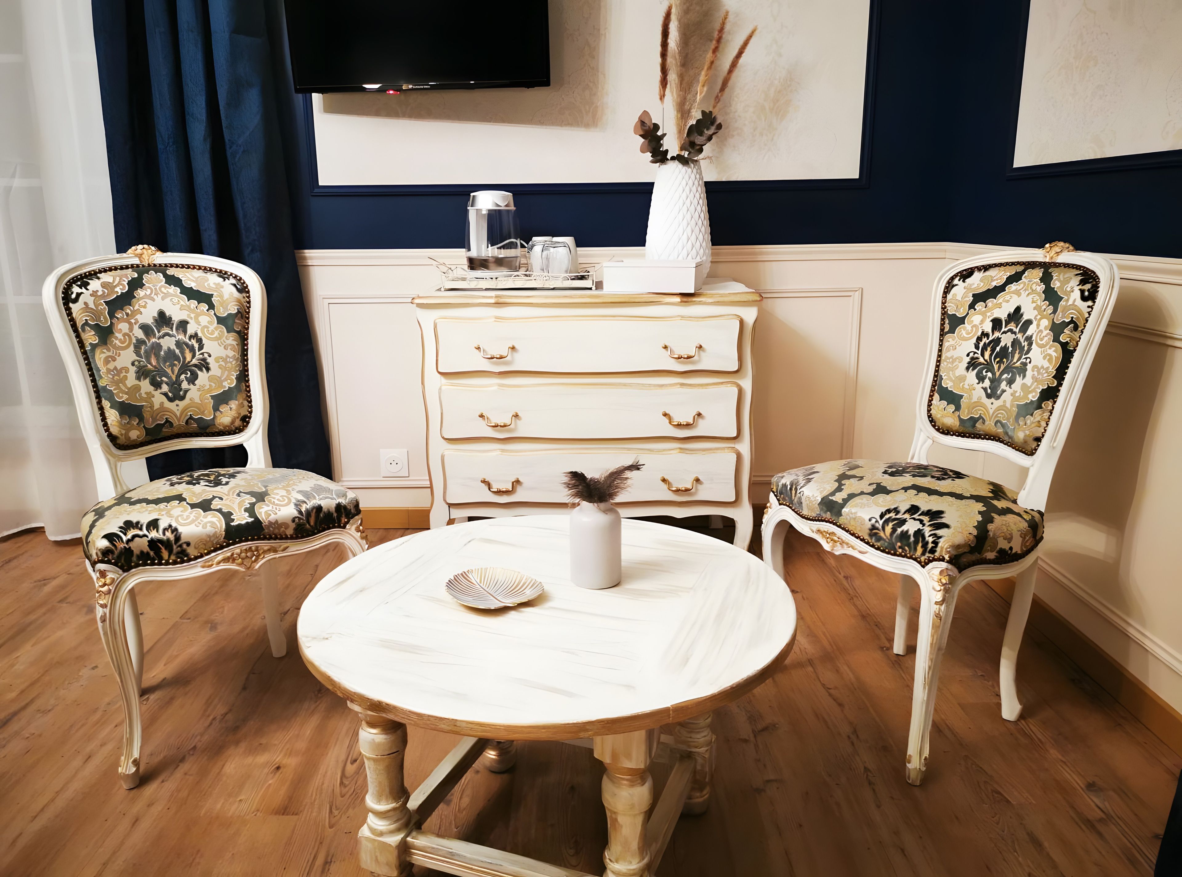 A room with two ornate chairs, a round wooden table, a white dresser with drawers, and a vase with dried plants on the dresser.