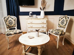 Una habitación con dos sillas ornamentadas, una mesa redonda de madera, un aparador blanco con cajones y un florero con plantas secas sobre el aparador.