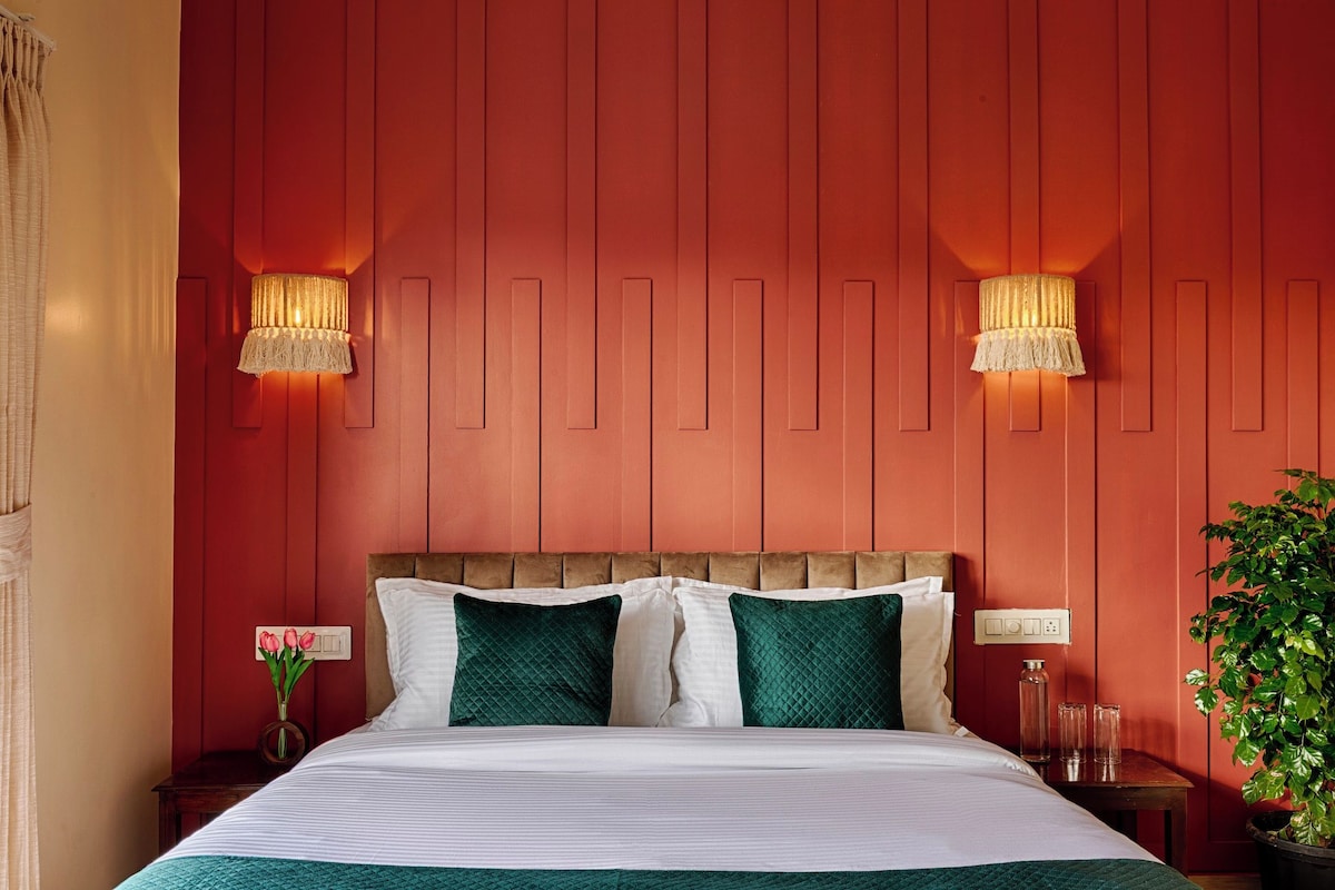 Hotellihuone, jossa on sänky, kaksi seinälle asennettua lamppua, yöpöytä, maljakko kukkineen ja ruukkukasvi.