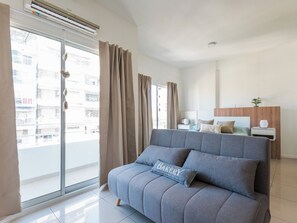 A modern bedroom with a grey sofa, a wooden headboard, a bedside table with a lamp, and a view of a residential area through the window. - Bright and comfortable apartment in Congreso (Buenos Aires)