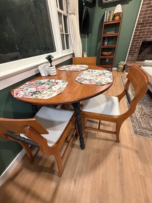 Una mesa de comedor con manteles individuales estampados con flores, sillas de madera y una ventana con cortinas.