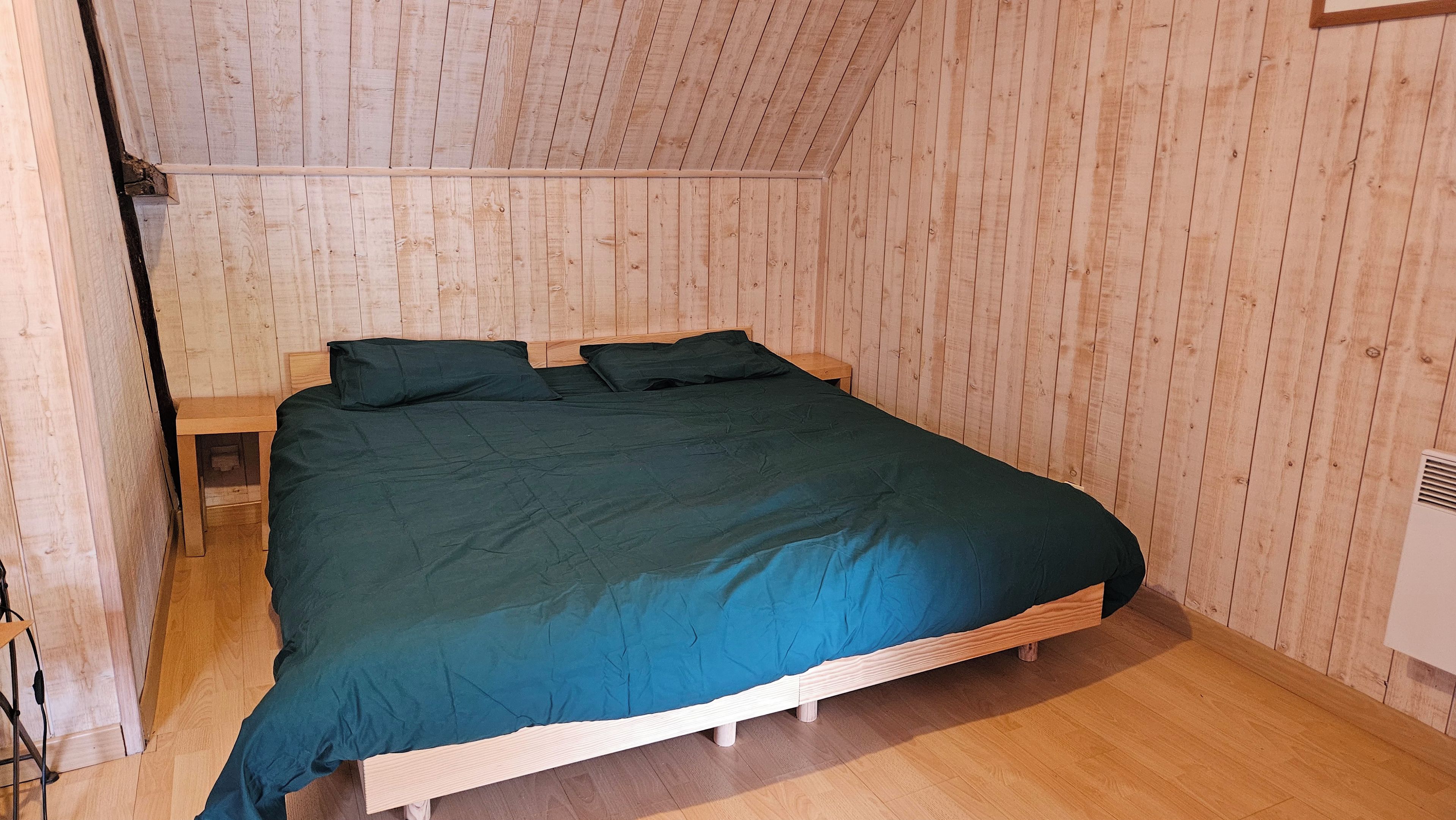 Un lit simple recouvert d’une couvre-lit vert foncé, dans une pièce aux murs et au plancher en bois.