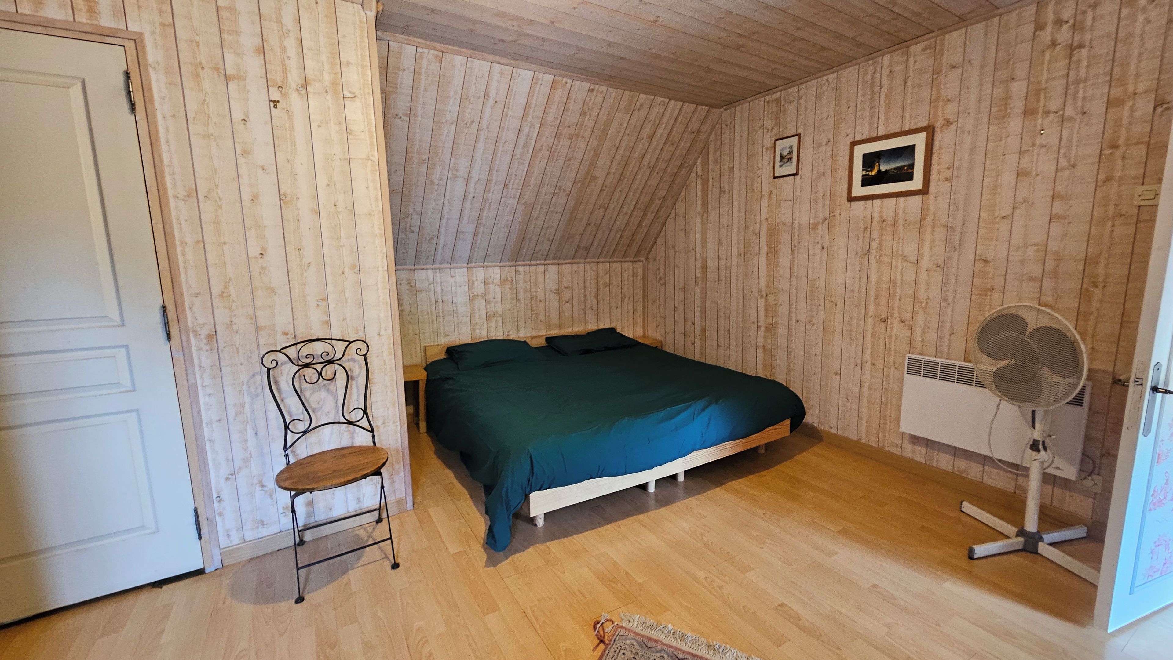 Een kamer met een schuine kap, een bed met een groene sprei, een stoel, een ventilator en een deur.
