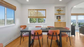 Un espace repas comprenant une grande table, six chaises, une fenêtre donnant sur un beau paysage, une étagère avec des objets décoratifs et une porte menant à un espace extérieur.