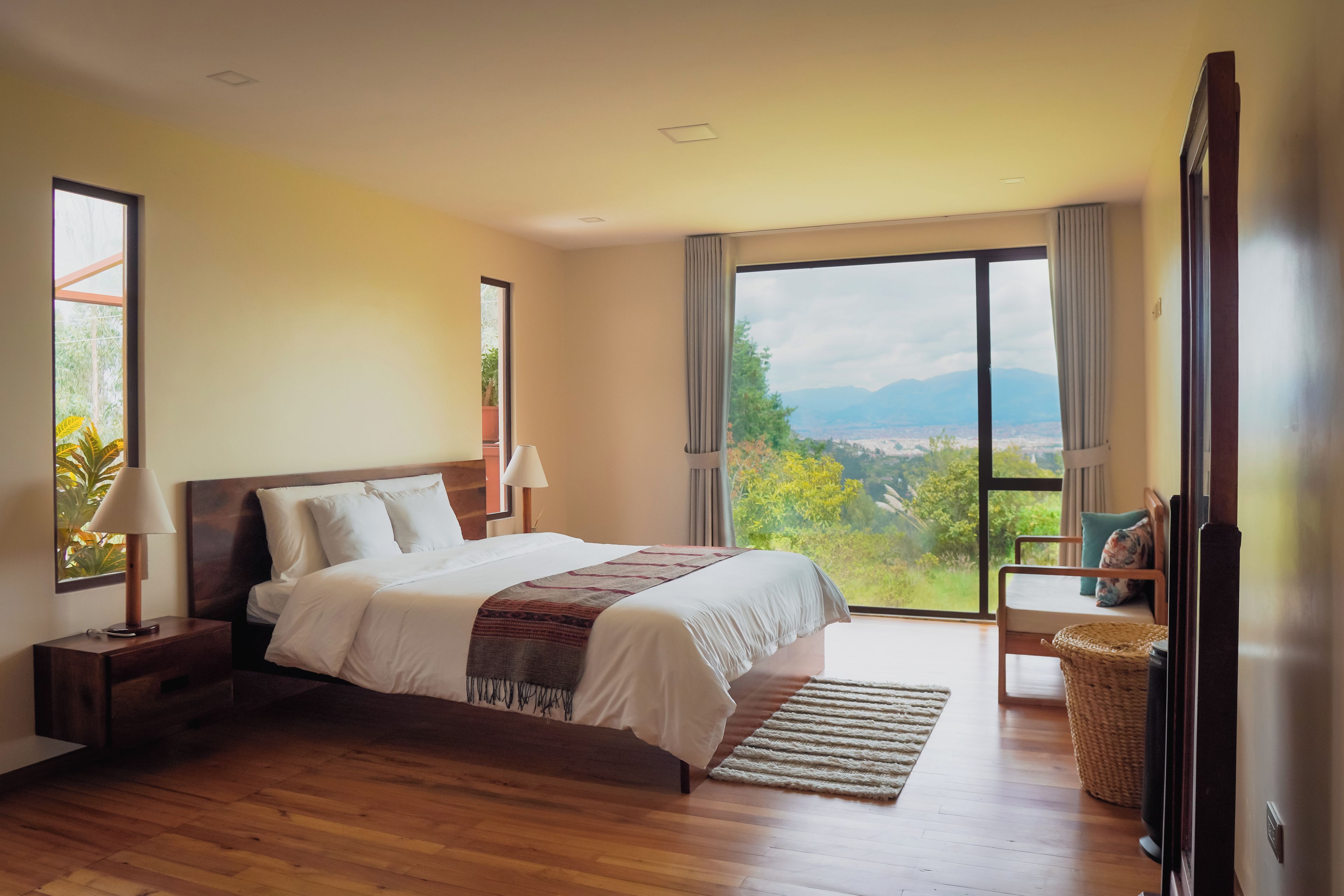 Ein Schlafzimmer mit einem großen Bett, einem Nachttisch, einer Lampe, einem Sessel und Blick ins Freie.
