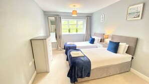 Quarto de hotel com duas camas, cômoda, espelho, janela com cortinas, luminária e um quadro na parede.