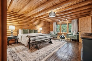 Ein Schlafzimmer in einer Blockhütte mit einem Bett, zwei Nachttischen, einer Bank, einem Sessel und einem Fenster mit Vorhängen.