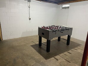 A room with a foosball table, white brick walls, and a concrete floor. - Remote River Retreat, Kayak, Fish, Canoe & More (Augusta)