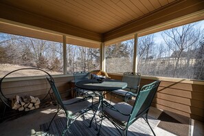 Une terrasse couverte avec une table ronde et des chaises, une vue sur les arbres et une pile de bois de chauffage.