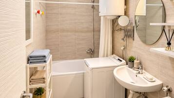 Een moderne badkamer met een badkuip, een witte wastafel en een toilet. Er is een plank met handdoeken en een klein plantje.