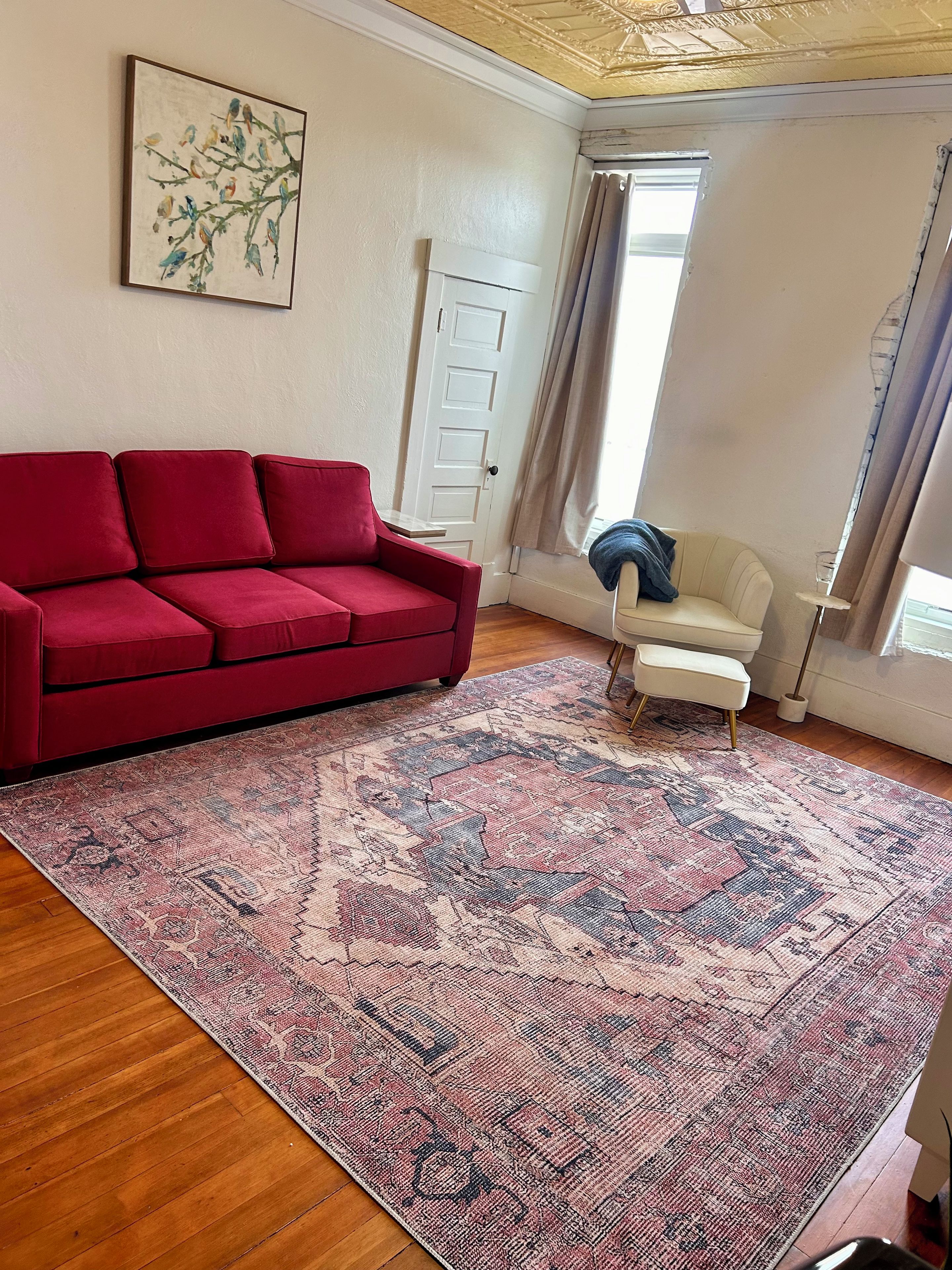 Una sala de estar con un sofá rojo, un sillón blanco, un tapete estampado y un cuadro de pájaros en la pared.