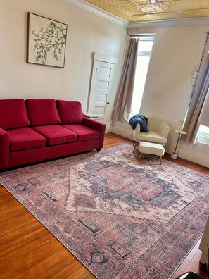 Una sala de estar con un sofá rojo, un sillón blanco, un tapete estampado y un cuadro de pájaros en la pared.
