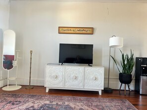 Una sala de estar con un mueble blanco, un televisor de pantalla plana, una alfombra estampada, una planta en maceta y un cuadro colgado en la pared.