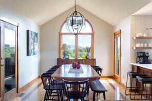 Un espace repas avec une table et des chaises en bois, un luminaire suspendu et une vue sur la verdure grâce à une grande fenêtre.