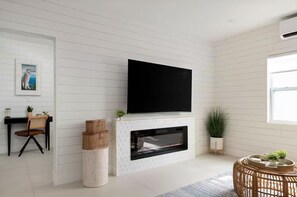 Een moderne woonkamer met een open haard, een flatscreen televisie erboven en een eethoek met een tafel en stoelen, en een groot raam dat voor natuurlijk licht zorgt.