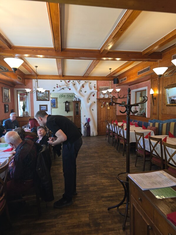 L’intérieur d’un restaurant est agrémenté de poutres en bois, de tables recouvertes de nappes blanches et d’un menu affiché sur un panneau mural.