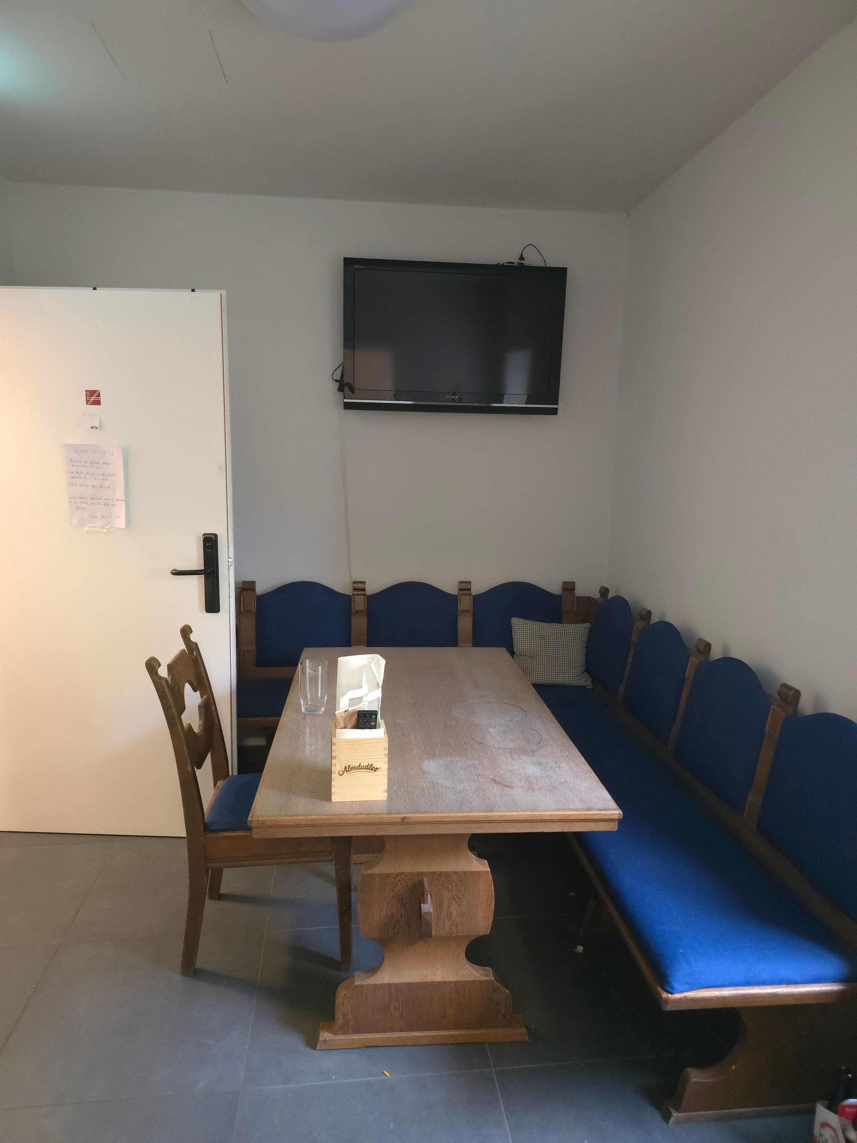 A room with a wooden table, a blue sofa, a television mounted on the wall, and a door with a notice on it.