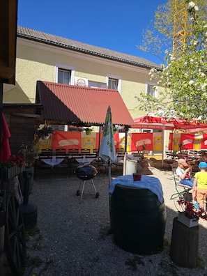Un espace extérieur aménagé avec des tables et des chaises, un barbecue et un bâtiment avec un toit rouge en arrière-plan.