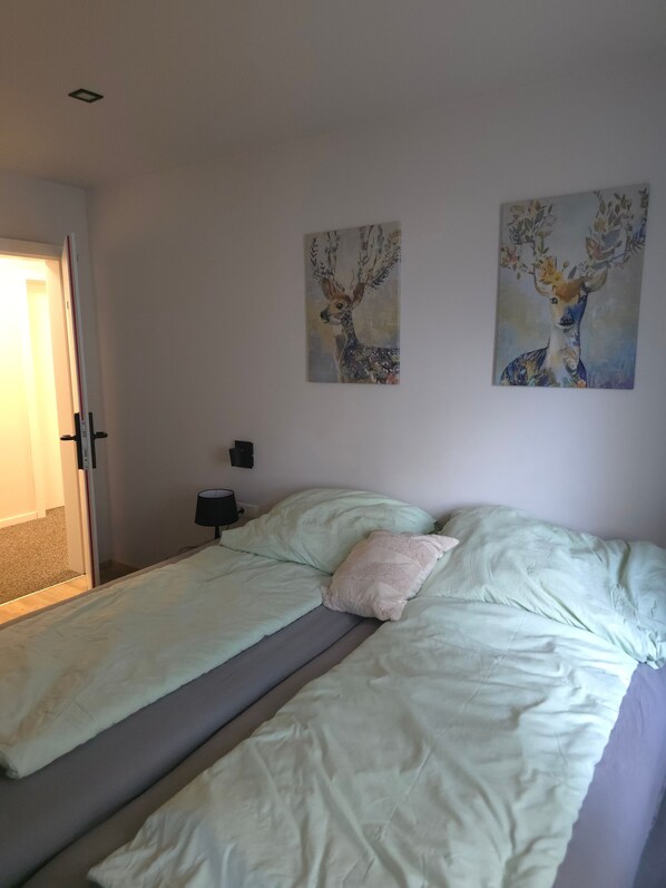 Une chambre à coucher avec un lit, deux tableaux accrochés au mur, une table de chevet avec une lampe, et une porte donnant sur une autre pièce.