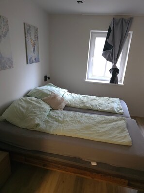 Un lit simple avec un matelas et des oreillers, une fenêtre avec des rideaux et deux tableaux accrochés au mur.