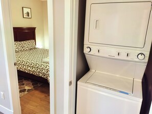 Un appareil compact combinant lave-linge et sèche-linge, placé dans une chambre à coucher, avec une couvre-lit à motifs et une tête de lit en bois.