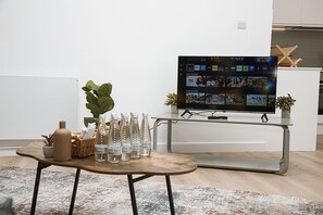 Een moderne woonkamer met een flatscreen televisie waarop verschillende zenders worden getoond, een bijzettafeltje met flessen en een vaas, en een potplant.