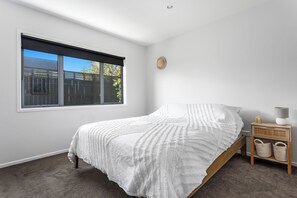 A bedroom with a bed, a window, a nightstand, and a lamp. - Coastal Calm - Ohope Beach Holiday Home (Ohope)