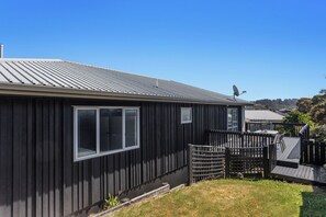 A single-story building with a dark exterior, a visible window, and a deck area with outdoor furniture. - Coastal Calm - Ohope Beach Holiday Home (Ohope)