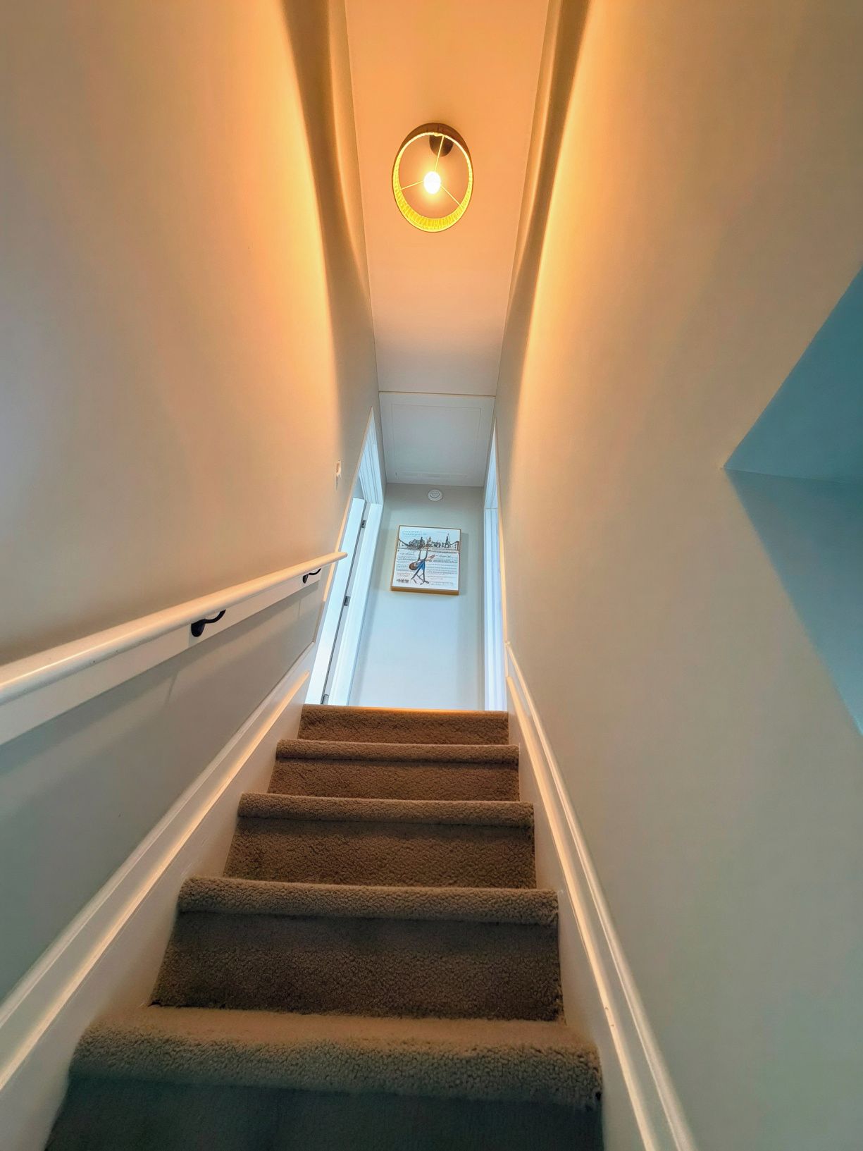 Una escalera alfombrada con barandilla, una lámpara en la parte superior y un cartel informativo enrabado en la parte inferior.