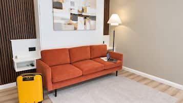 A room with a sofa, a lamp, a small table, a suitcase, and a painting on the wall.