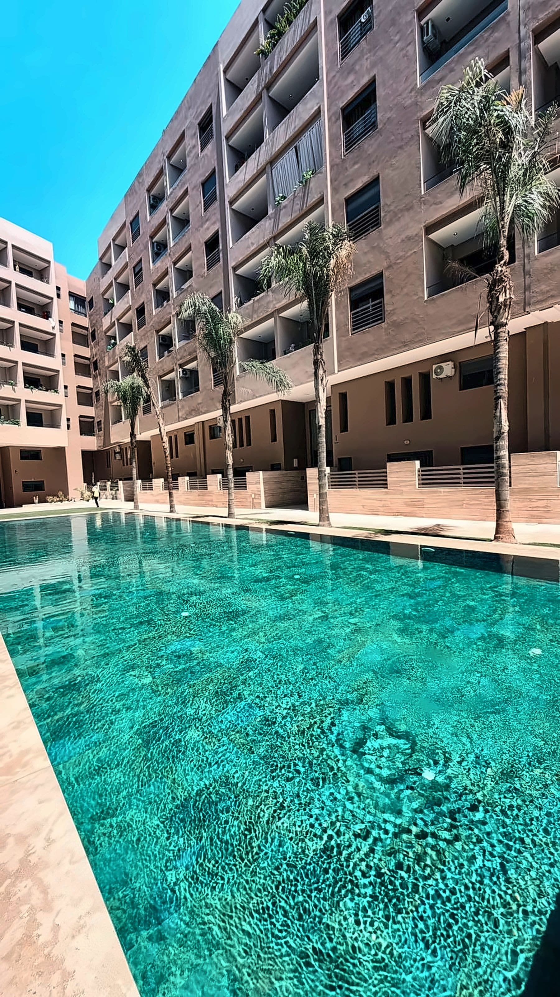 A swimming pool with clear water, surrounded by a multi-story building with balconies and palm trees.