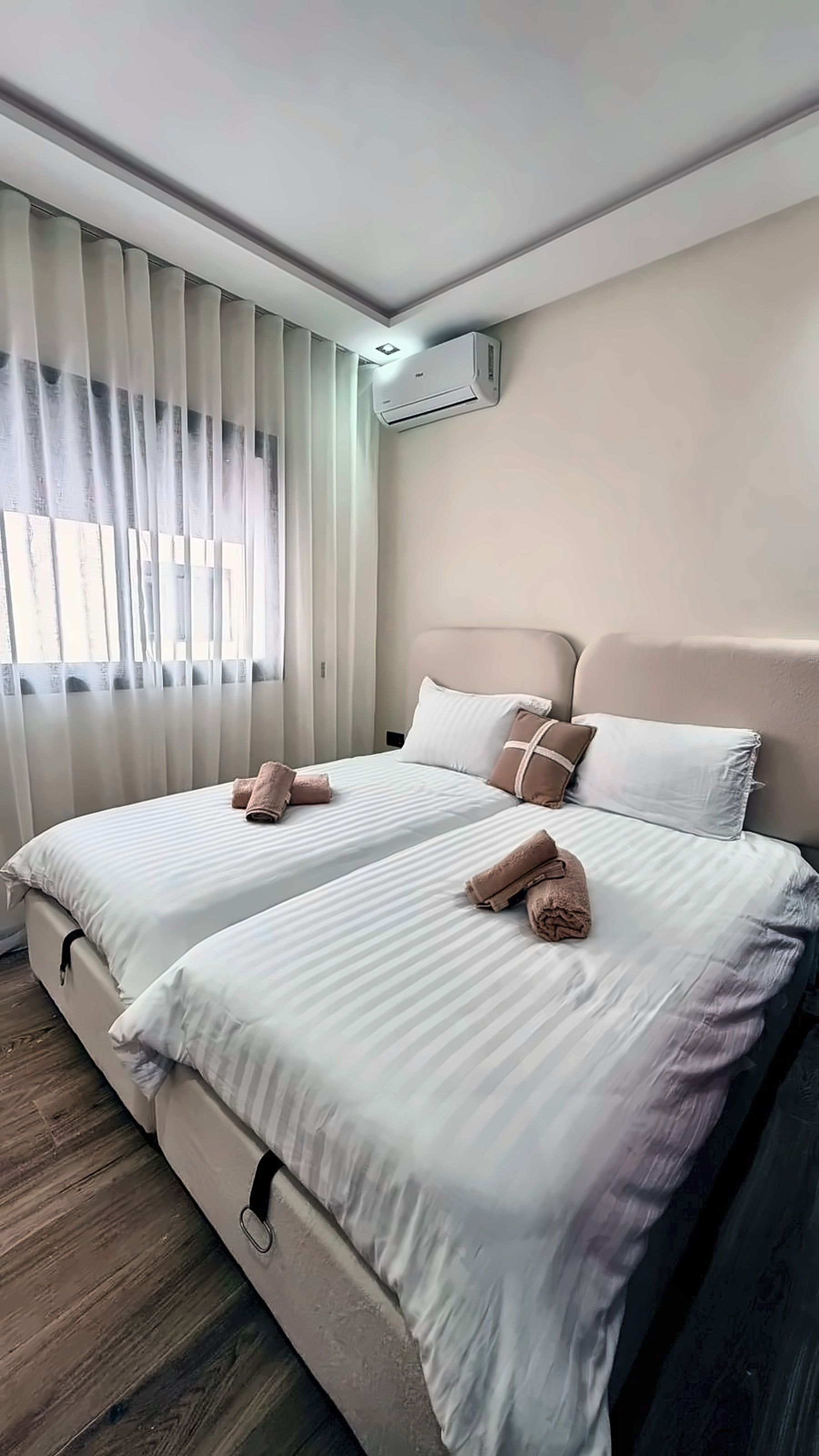 A hotel room with two beds, a ceiling-mounted air conditioner, and a window with curtains.