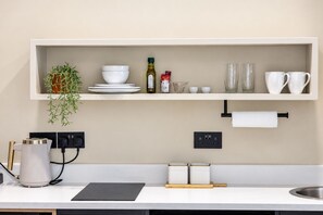 Une cuisine moderne avec un plan de travail blanc, une étagère intégrée où sont rangés de la vaisselle, une plante et une bouilloire.