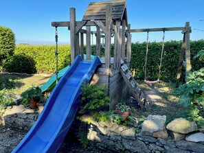 Une structure de jeu en bois, dotée d’un toboggan et de balançoires, installée dans un jardin.