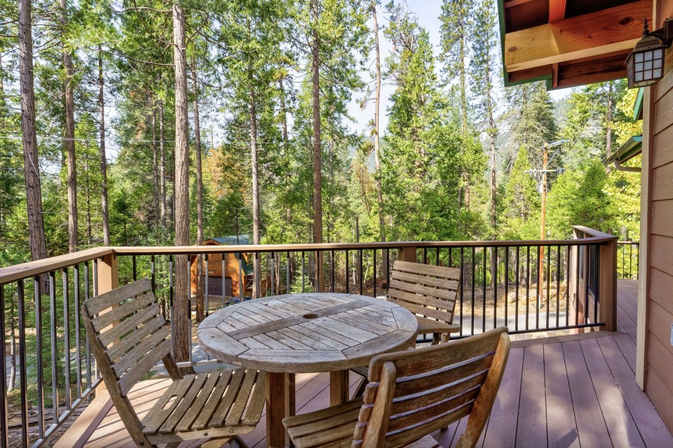 A wooden deck with a round table and chairs, surrounded by a metal railing, overlooking a forested area.